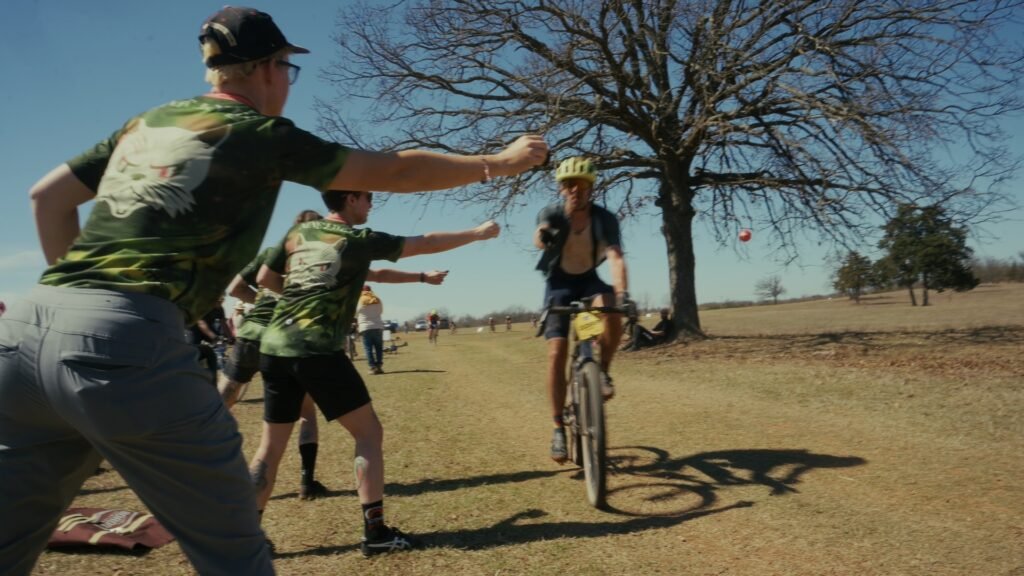 Three members of Nine Lives Cycling Collective in matching team shirts give gummy bear hand ups to racers coming through the Bovine Bypass Aid Station at the MidSouth 2026.