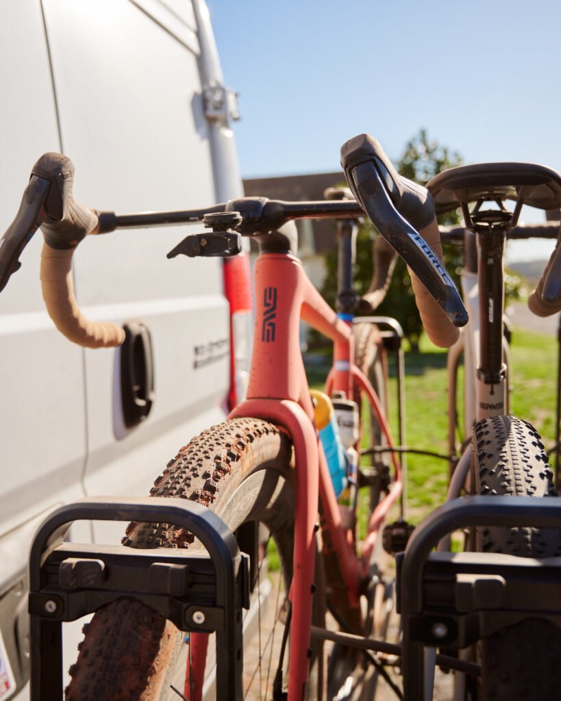 A wayfinder coral ENVE MOG sits on a Kuat Piston Pro bike rack behind a white van.