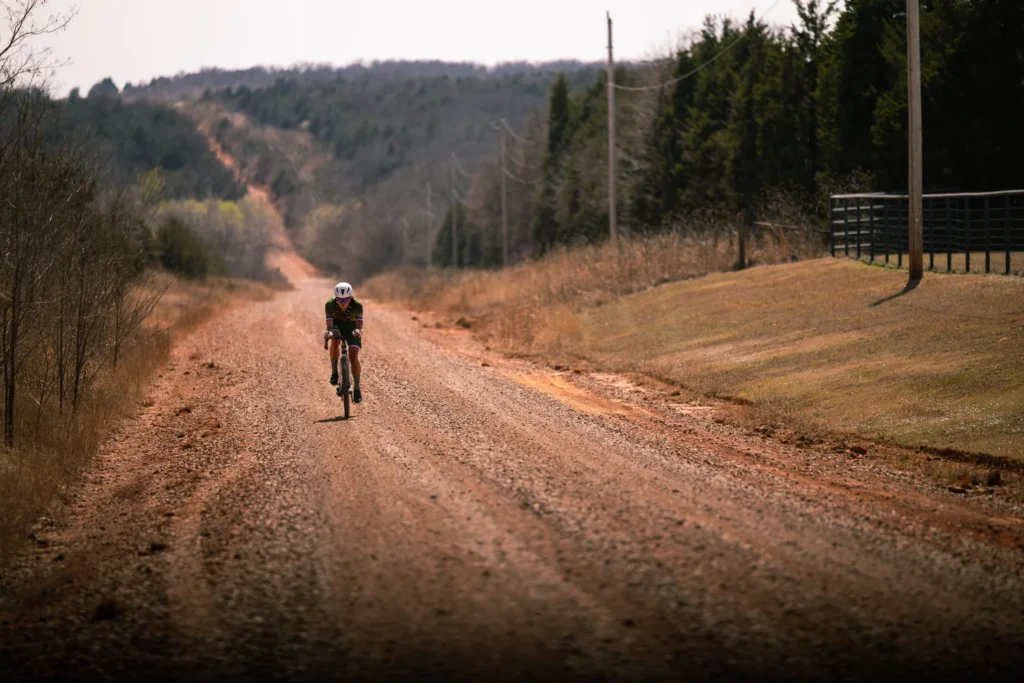 Robin Cummings rides a gravel bike solo in a Nine Lives Cycling skinsuit on an empty red dirt road in Stillwater, OK